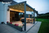 Pergola przyścienna Elegancy Tarasola o szerokości 6 metrów, z elektrycznie rozsuwanym dachem tkaninowym i wbudowanym oświetleniem LED, tworzy elegancką strefę jadalnianą na nowoczesnym tarasie. Konstrukcja harmonizuje z architekturą domu, a w tle widoczny jest zadbany trawnik i roślinność.