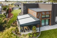 Wolnostojąca pergola ogrodowa Elegancy Tarasola o imponującej szerokości 9 metrów tworzy dużą przestrzeń wypoczynkową z sofami i stolikami, otoczoną nowoczesnym ogrodem i bujną roślinnością. Widok z góry podkreśla skalę konstrukcji oraz jej harmonijne dopasowanie do krajobrazu.