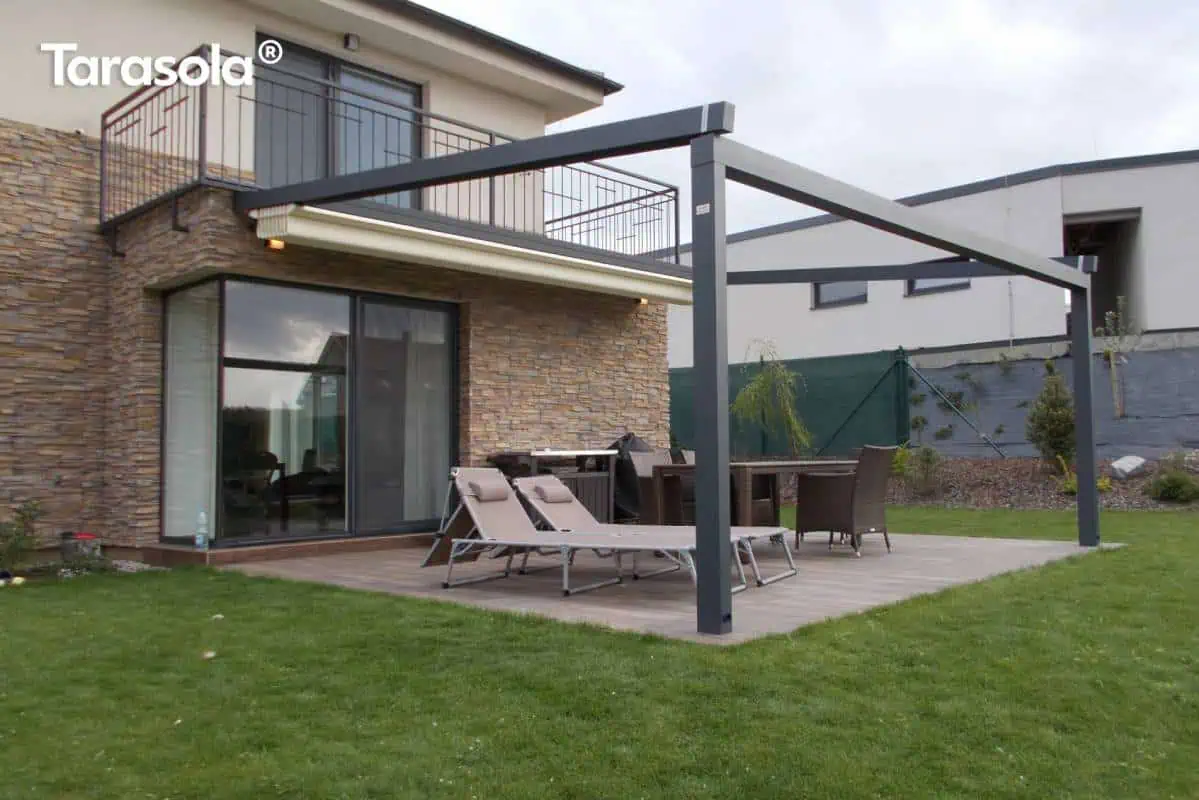 Pergola ścienna Elegancy Tarasola o minimalistycznej, aluminiowej konstrukcji tworzy zadaszoną przestrzeń na tarasie z zestawem wypoczynkowym i leżakami. Solidne profile podkreślają nowoczesny styl aranżacji, a wokół znajduje się ogród oraz sąsiednie zabudowania.