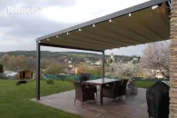 Pergola tarasowa Elegancy Tarasola z aluminiową konstrukcją na dwóch podporach i tkaninowym dachem tworzy komfortową strefę jadalnianą na dużym tarasie. Jasna tkanina dodaje lekkości aranżacji, a w tle rozciąga się widok na zielone pagórki, ogród oraz zabudowania w oddali.