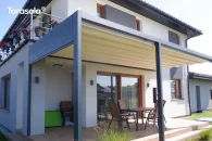 Tarasola Flat z aluminiową konstrukcją przyścienną oraz rozsuwanym, kremowym dachem tkaninowym tworzy elegancką zabudowę tarasu przy domu. Stabilne podpory w grafitowym kolorze podkreślają minimalistyczny wygląd pergoli, a pod nią ustawiono drewniany stół z krzesłami. Otoczenie stanowi zadbany ogród i nowoczesna architektura.