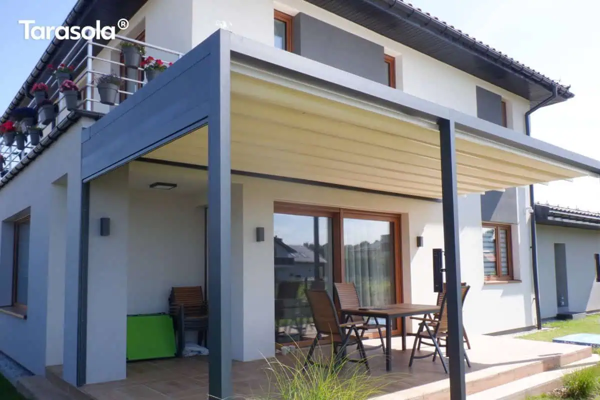 Aluminiowa zabudowa tarasu z pergolą Flat Tarasola Tarasola Flat z aluminiową konstrukcją przyścienną oraz rozsuwanym, kremowym dachem tkaninowym tworzy elegancką zabudowę tarasu przy domu. Stabilne podpory w grafitowym kolorze podkreślają minimalistyczny wygląd pergoli, a pod nią ustawiono drewniany stół z krzesłami. Otoczenie stanowi zadbany ogród i nowoczesna architektura.