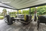 Pergola Elegancy Tarasola z rozciąganym dachem tkaninowym osłania przestrzeń wypoczynkową z wygodnymi fotelami i sofami w nowoczesnym, grafitowym stylu. Zadaszenie idealnie komponuje się z dużym tarasem wyłożonym płytami, otoczonym ogrodem pełnym zieleni.