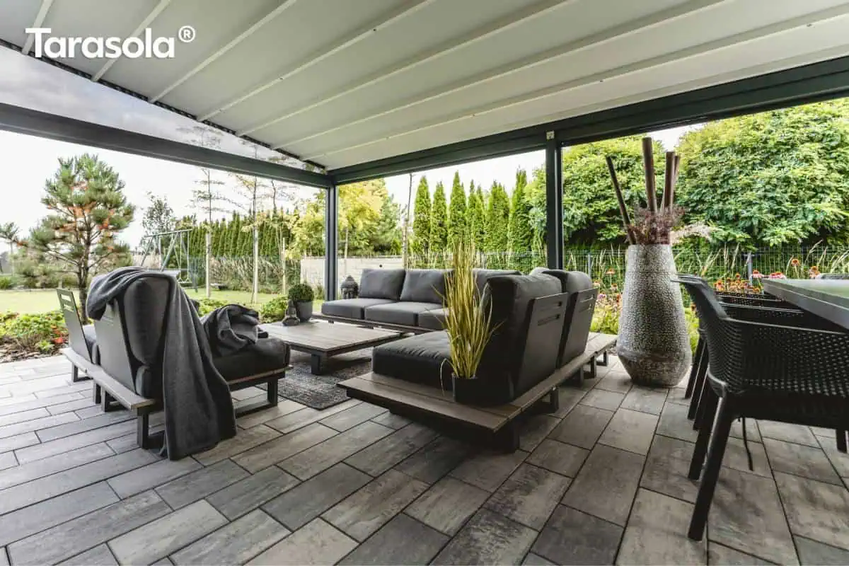 Pergola Elegancy Tarasola z rozciąganym dachem tkaninowym osłania przestrzeń wypoczynkową z wygodnymi fotelami i sofami w nowoczesnym, grafitowym stylu. Zadaszenie idealnie komponuje się z dużym tarasem wyłożonym płytami, otoczonym ogrodem pełnym zieleni.