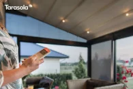 Pergola Elegancy Tarasola z elektrycznie rozsuwanym dachem i zintegrowanym oświetleniem LED tworzy przyjemną, wieczorną atmosferę na tarasie. Na pierwszym planie osoba obsługuje pergolę za pomocą pilota, a w tle widoczny jest ogród oraz nowoczesne meble tarasowe.