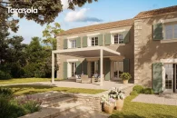 Pergola bioklimatyczna z elektrycznie obracanymi lamelami i dwoma przednimi podporami, w jasnym kremowo-białym kolorze, zamontowana przy elewacji domu; tworzy nowoczesne zadaszenie tarasu z zestawem wypoczynkowym w otoczeniu kamiennej nawierzchni i ogrodu pełnego roślin ozdobnych.