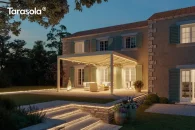 Przestronna pergola bioklimatyczna w kremowo-białym kolorze, z dużym modułem obracanych lameli sterowanych elektrycznie; tworzy elegancką strefę wypoczynkową na tarasie kamiennym przed klasycznym domem z okiennicami, otoczonym bujną zielenią ogrodu.