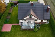 Duża pergola lamelowa w wersji przyściennej, w jasnym wykończeniu kremowo-białym, z elektrycznym sterowaniem lamelami; tworzy zacienioną strefę relaksu przed fasadą kamiennego domu z klasycznymi okiennicami i zielonym, zadbanym ogrodem.