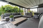 Aluminiowa pergola Elegancy Tarasola z rozciąganym elektrycznie dachem tkaninowym tworzy przestronną, nowoczesną strefę wypoczynkową z kompletem grafitowych sof i stolików. Konstrukcja osłania taras od góry i boków, zapewniając przytulny klimat w otoczeniu zadbanego ogrodu z bujną zielenią.