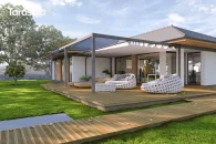 Aluminiowa pergola wolnostojąca Tarasola Flat z elektrycznie rozsuwaną tkaniną dachu oraz pełnym obwodem aluminiowych podpór tworzy duże zadaszenie nad tarasem. Konstrukcja osłania drewniany taras z designerskimi, okrągłymi fotelami, otoczony szerokim, zadbanym trawnikiem i nowoczesną zabudową domu.