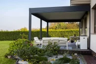 Wielkogabarytowa pergola przyścienna w kolorze antracytowym z lamelowym dachem sterowanym elektrycznie; pod zadaszeniem znajduje się jasny zestaw wypoczynkowy, otoczony bujnym ogrodem z roślinami ozdobnymi i dekoracyjną rabatą.