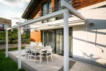 Lekka konstrukcja pergoli Tarasola Simple mocowanej do ściany, z cienkimi profilami wspornikowymi oraz prowadnicami przeznaczonymi do obsługi tkaninowego dachu. Pergola osłania taras jadalniany, a w otoczeniu widać ogrodowe rośliny i zabudowę sąsiednich domów.