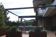 Pergola Tarasola Flat o prostokątnej konstrukcji przyściennej z całkowicie zebranym pakietem tkaniny dachu, eksponująca elegancką formę aluminiowych profili z rynną odpływową po lewej stronie konstrukcji. Zadaszenie osłania przestrzeń wypoczynkową z sofą i donicami dekoracyjnymi na tarasie otoczonym zielenią.