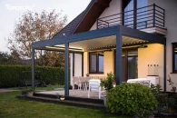 Pergola bioklimatyczna montowana do ściany, w antracytowym kolorze, z obróconymi lamelami tworzącymi cień; elegancko wykończony taras z białymi meblami ogrodowymi otacza zielony trawnik i roślinność ogrodowa, tworząc letnią, relaksującą atmosferę.