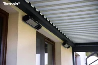 Bioklimatyczna pergola przyścienna z obracanymi lamelami dachowymi w antracytowej konstrukcji, pod pergolą widoczny minimalistyczny układ promienników ciepła.