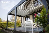 Rozległa konstrukcja bioklimatyczna z obracanymi lamelami, zamocowana do ściany domu w antracytowym kolorze. Na zdjęciu pergola tworzy komfortową strefę wypoczynku pod solidnym zadaszeniem, a pod nią znajdują się jasne meble ogrodowe otoczone bujną, zadbaną roślinnością.