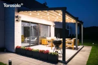 Pergola Elegancy Tarasola prezentuje system LED rozmieszczony w dachu tkaninowym, który podkreśla nowoczesny styl zadaszenia tarasowego. Oświetlona konstrukcja otacza strefę wypoczynku z drewnianymi meblami, a tło stanowi przestronny trawnik.
