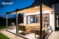 Taras z pergolą Elegancy Tarasola na trzech nogach aluminiowych, wyposażoną w równomierne oświetlenie punktowe LED, tworzy przyjemną, wieczorną strefę do relaksu przy drewnianych meblach ogrodowych. Wokół rozciąga się trawnik oraz spokojna zabudowa jednorodzinna.