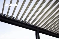 Otwarte pióra lameli dachu Tarasola Essential ukazują bioklimatyczne działanie pergoli, której lamele ustawione są tak, aby wpuszczać światło i świeże powietrze. Konstrukcja w antracycie kontrastuje z jasnymi lamelami, a w tle widoczny jest delikatnie zachmurzony, jasny nieboskłon.