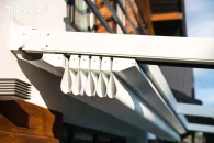 Zbliżenie na pakiet złożonej tkaniny dachu pergoli Tarasola Simple mocowanej do ściany, prezentujące lekką konstrukcję i system zwijania poszycia, które układa się w dekoracyjne fale. W tle widoczna fasada domu i elementy ogrodowej zabudowy.