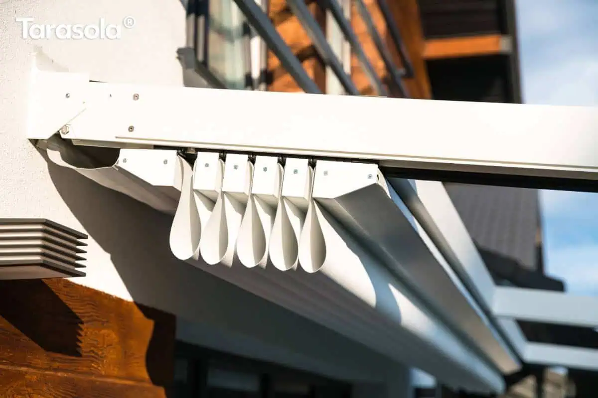 Zbliżenie na pakiet złożonej tkaniny dachu pergoli Tarasola Simple mocowanej do ściany, prezentujące lekką konstrukcję i system zwijania poszycia, które układa się w dekoracyjne fale. W tle widoczna fasada domu i elementy ogrodowej zabudowy.