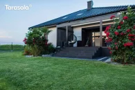 Pergola Elegancy Tarasola zamontowana nad wejściem do domu tworzy nowoczesne, aluminiowe zadaszenie z tkaninowym dachem, osłaniając strefę wypoczynkową z wiszącym fotelem. Konstrukcja harmonizuje z elewacją i otaczającym ogrodem pełnym roślin oraz krzewów róż.