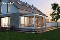 Elegancka biała pergola Elegancy Tarasola z pełnymi szklanymi drzwiami tworzy elegancką, półzamkniętą przestrzeń wypoczynkową oświetloną subtelnymi punktami LED. Zabudowa osłania stylowe meble tarasowe, a jasny dom i ogród z rabatami nadają aranżacji nowoczesny charakter.