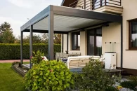 Pergola Tarasola Essential z trwałą konstrukcją przyścienną nad eleganckim tarasem z jasnymi meblami ogrodowymi i zadbaną roślinnością w ogrodzie