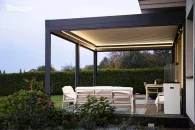 Lamelowa osłona tarasowa Tarasola Essential zamocowana do elewacji domu prezentuje solidną, antracytową konstrukcję podkreśloną delikatnym oświetleniem LED, a wokół niej rozciąga się zielony ogród, którego rośliny tworzą spokojny, naturalny klimat.