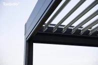 W pergoli Tarasola Essential uwagę przyciąga roleta boczna, która domyka przestrzeń wypoczynkową i chroni ją przed podmuchami wiatru, a jasna aranżacja mebli ogrodowych pięknie komponuje się z zielenią żywopłotu i miękkim światłem popołudnia.