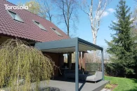 Pergola Tarasola Flat o prostokątnej, aluminiowej konstrukcji montowanej przy ścianie domu, z jasnym dachem tkaninowym rozwijanym w prowadnicach i punktowym oświetleniem LED. Zadasza taras z narożną sofą ogrodową, otoczony zielonym ogrodem, drzewami i trawnikiem.