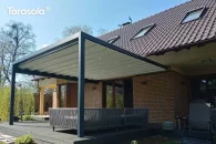Duża pergola Tarasola Flat w kolorze antracytowym, zamontowana przy ścianie budynku, z szerokim, zwijanym dachem tkaninowym i solidnymi aluminiowymi profilami. Osłania przestronny taras wypoczynkowy z sofami, położony w otoczeniu ogrodu z wysokimi drzewami i krzewami.