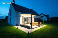 Pergola ogrodowa Elegancy Tarasola z podświetlanym dachem tkaninowym, wsparta na trzech przednich podporach, tworzy stylową przestrzeń wypoczynkową przy domu, z drewnianymi meblami tarasowymi o jasnych poduszkach. Zadaszenie subtelnie rozświetla ogród i sąsiednie budynki w letni wieczór.