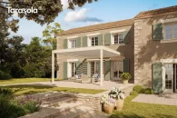 Biała pergola przyścienna Essential Tarasola z regulowanym dachem lamelowym tworzy elegancką, zadaszoną strefę jadalnianą, a otaczają ją geometryczne rabaty, niska zieleń i ciepła kolorystyka elewacji, które razem budują harmonijny tarasowy klimat.