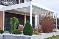 Biała, wolnostojąca pergola Tarasola Flat z tkaninowym, rzymskim dachem oraz wbudowanymi punktami LED oświetla tarasową strefę relaksu. Prosta, aluminiowa konstrukcja stoi w ogrodzie pełnym ozdobnych roślin i nowoczesnych nasadzeń.