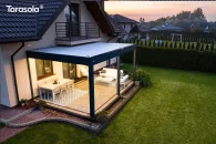 Pergola Tarasola Essential zamontowana przy ścianie domu zapewnia cienistą przestrzeń do relaksu, a jasne meble i neutralne dywany wzbogacają aranżację tarasu, który sąsiaduje z dużym, zadbanym ogrodem pełnym zieleni.