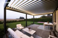 Regulowany dach przeciwsłoneczny pergoli Tarasola Essential tworzy komfortową osłonę nad zestawem wypoczynkowym, a szeroki widok na ogród, równo przycięte krzewy i otwartą przestrzeń podkreśla spokojny, relaksujący charakter tarasu.