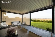 Nowoczesna pergola Essential Tarasola z lamelowym dachem i rozwiniętymi roletami wiatrochronnymi tworzy przytulną, osłoniętą strefę wypoczynkową na jasnym tarasie, z widokiem na zadbany, szeroki trawnik i eleganckie nasadzenia w tle.