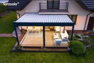 Aluminiowa pergola Essential od Tarasoli tworzy elegancką, dopasowaną do elewacji przestrzeń wypoczynkową z pełnym zadaszeniem z lameli, które skutecznie chronią taras. Całość otacza zadbany ogród i równy, zielony trawnik, które podkreślają uporządkowany charakter strefy relaksu.