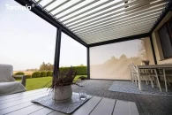 W pergoli Essential od Tarasoli półprzejrzysta roleta boczna delikatnie rozprasza światło i zapewnia prywatność, nie odbierając poczucia przestrzeni. Za osłoną rozciąga się zadbany ogród z geometrycznymi krzewami, wprowadzający spokojny, domowy charakter.