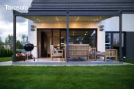Przyścienna pergola aluminiowa Elegancy Tarasola z oświetlanym dachem tkaninowym osłania elegancką strefę wypoczynkową na tarasie, wyposażoną w drewniane meble i grill. Podświetlenie tworzy ciepły klimat, a w tle widoczny jest ogród i nocne niebo.