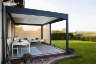 Pod zadaszeniem z obracanych lameli pergoli Tarasola Essential powstała nastrojowa, przytulna przestrzeń tarasowa z jasnym kompletem sof i krzeseł. Konstrukcja w antracycie harmonizuje z delikatnymi beżowymi roletami, a klimat miejsca budują dywany zewnętrzne, dekoracje stołowe i otulająca zieleń ogrodu widoczna za osłonami.