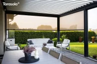 Pergola przyścienna Essential Tarasola wyposażona w roletę ZIP, która chroni taras przed wiatrem i niskim słońcem. Antracytowa konstrukcja z jasnymi lamelami dachowymi tworzy nowoczesny kontrast, a wnętrze z komfortowymi meblami wypoczynkowymi otwiera się na starannie utrzymany ogród z żywopłotem i rabatami roślinnymi.