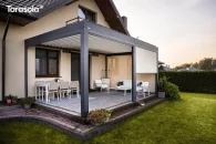 Zabudowana roletami tarasowymi pergola Essential, której lamelowy dach i boczne przesłony tworzą półprywatną, elegancką strefę wypoczynkową. Antracytowe profile oraz beżowe rolety wprowadzają nowoczesną stylistykę, a z tarasu roztacza się widok na zadbaną, zieloną przestrzeń ogrodu z żywopłotem i kwietnymi nasadzeniami.