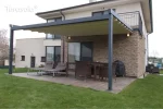 Pergola materiałowa Elegancy Tarasola z elektrycznie rozwijanym dachem tkaninowym osłania taras z leżakami oraz wiklinowym zestawem jadalnianym. Konstrukcja w grafitowym kolorze harmonizuje z elewacją domu z kamiennym wykończeniem, a otoczenie stanowi ogród z zadbanym trawnikiem.