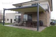 Pergola materiałowa Elegancy Tarasola z elektrycznie rozwijanym dachem tkaninowym osłania taras z leżakami oraz wiklinowym zestawem jadalnianym. Konstrukcja w grafitowym kolorze harmonizuje z elewacją domu z kamiennym wykończeniem, a otoczenie stanowi ogród z zadbanym trawnikiem.