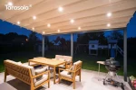 Ścienna pergola rzymska Elegancy Tarasola z podświetlanym dachem tkaninowym tworzy przyjemną, wieczorną strefę wypoczynkową z drewnianymi meblami i grillem. Zadaszenie rozświetla taras, a w tle widoczne są ogród, plac zabaw i zabudowa jednorodzinna.