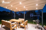 Ścienna pergola rzymska Elegancy Tarasola z podświetlanym dachem tkaninowym tworzy przyjemną, wieczorną strefę wypoczynkową z drewnianymi meblami i grillem. Zadaszenie rozświetla taras, a w tle widoczne są ogród, plac zabaw i zabudowa jednorodzinna.