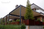 Pergola Tarasola Flat zamontowana do elewacji, wyposażona w ruchomy dach tkaninowy z wbudowanym oświetleniem LED i prostokątną konstrukcję aluminiową. Osłania elegancki taras z dużymi donicami i roślinnością ozdobną, w otoczeniu nowoczesnej architektury i ogrodowej zieleni.