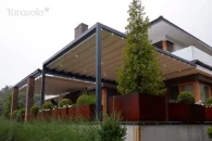 Pergola Tarasola Flat zamontowana do elewacji, wyposażona w ruchomy dach tkaninowy z wbudowanym oświetleniem LED i prostokątną konstrukcję aluminiową. Osłania elegancki taras z dużymi donicami i roślinnością ozdobną, w otoczeniu nowoczesnej architektury i ogrodowej zieleni.